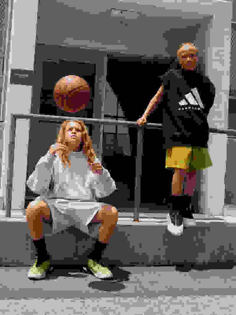 A kid sits, throwing a basketball, next to a kid leaning on a railing