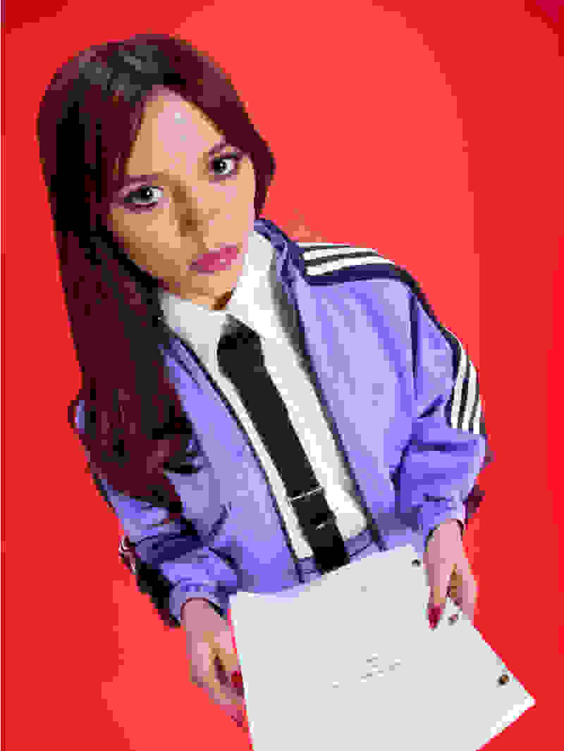 Actress Jenna Ortega, dressed in purple adidas tracksuit and neck tie, holds a script in hand.