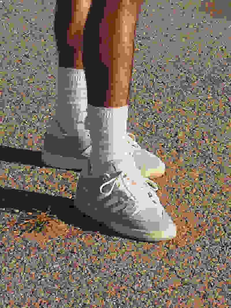 A knee-down view of a man standing on pavement, wearing white crew socks and grey adidas Forum sneakers.
