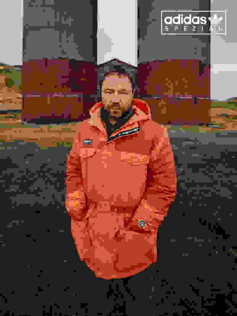 Stephen Graham stands firmly in an industrial landscape, looking sternly into the camera while wearing an orange adidas Spezial jacket