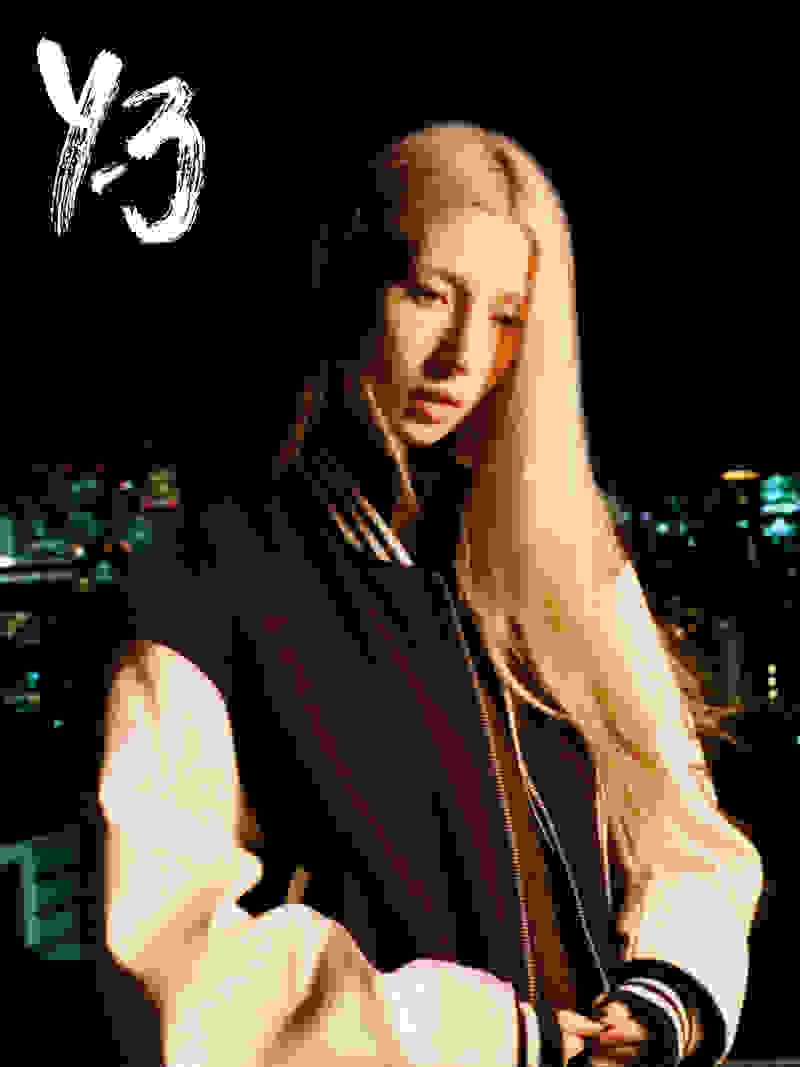 A blonde woman wearing a black and white Y-3 jacket poses in front of a dark sky with city lights visible in the background.