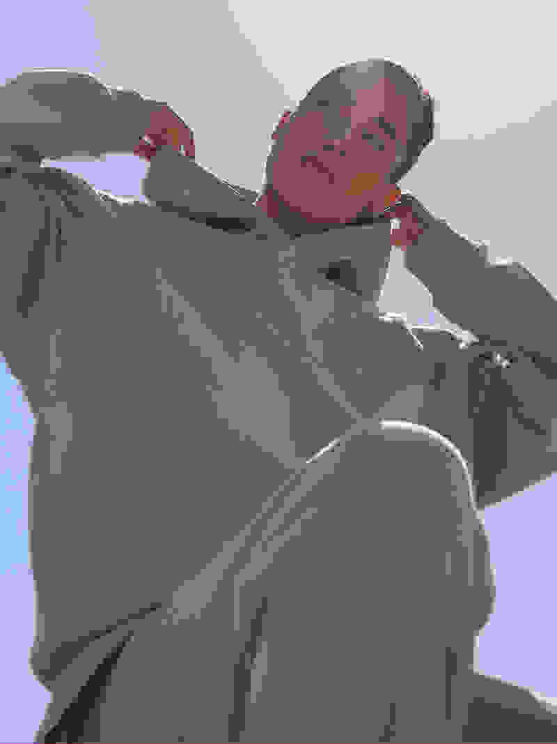 A "ground up" view of a woman crouched wearing a grey sweatsuit and adjusting the hood with a bright, sunny sky in the background.