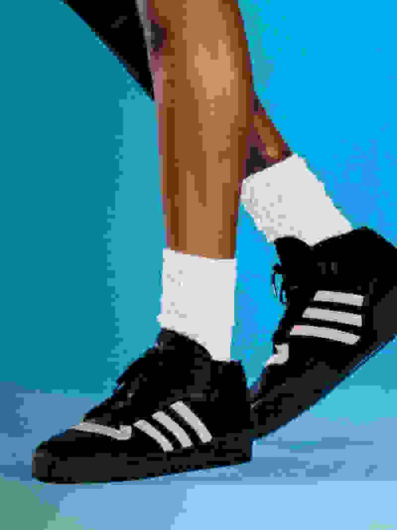 Close-up of model's legs wearing white socks and black adidas shoes with off-white stripes against blue background