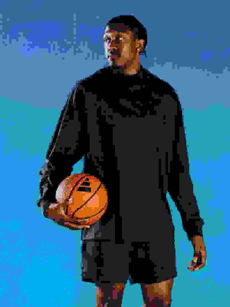 Male athlete wearing black top and shorts stands and holds basketball in front of blue background
