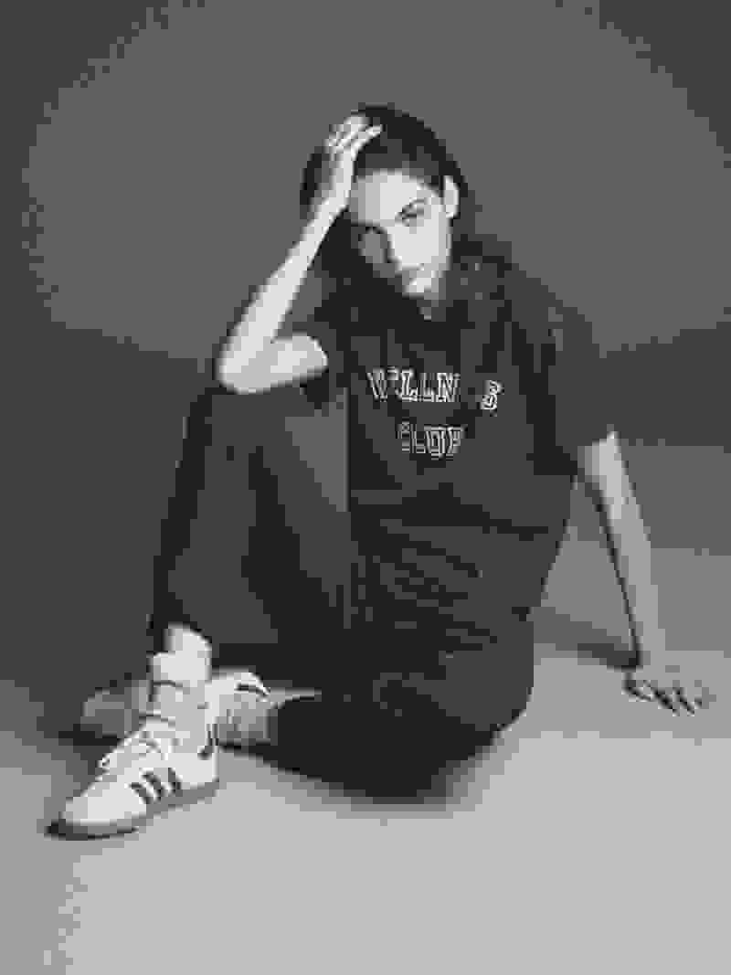 A woman with dark hair wearing jeans, a black tee, and the adidas Originals x Sporty & Rich Sambas sits on a white floor in an empty room.