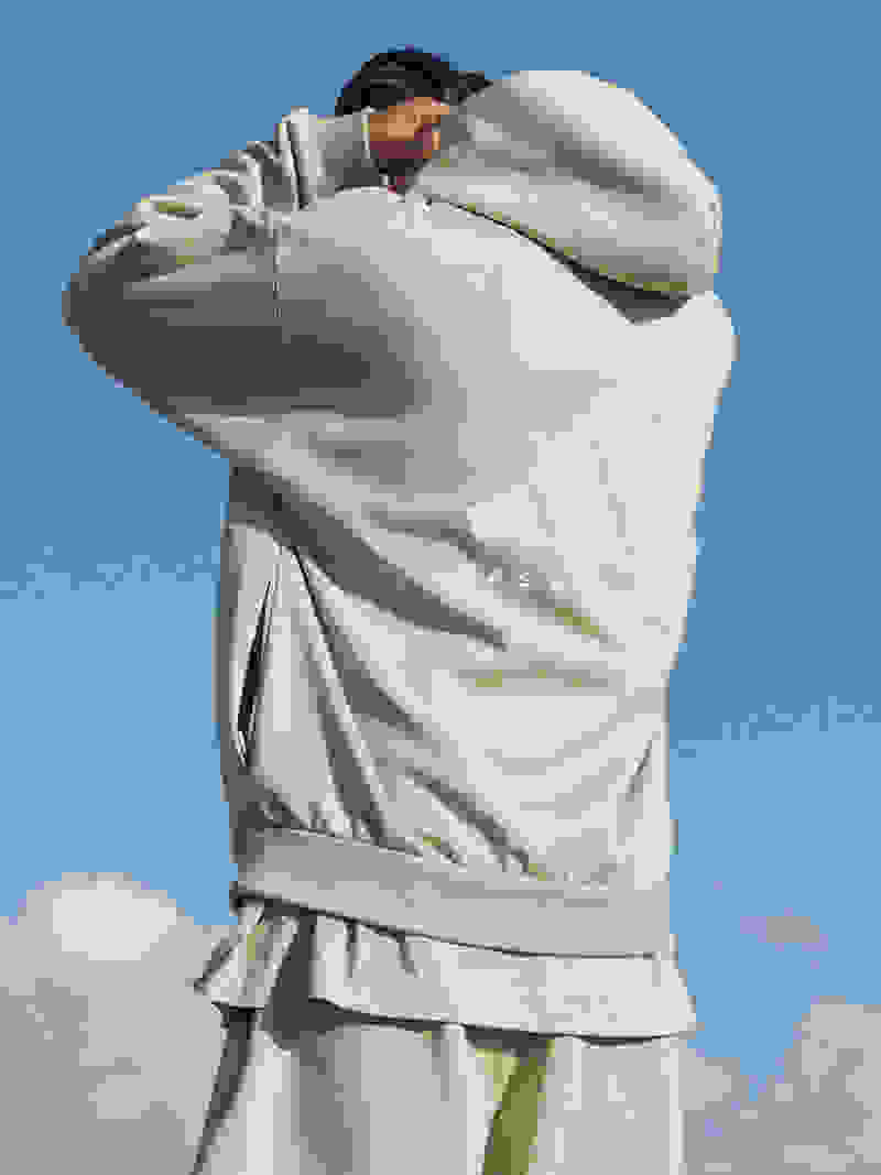 A rear view of a man standing under a blue sky, adjusting the hood of his pale green adidas sweatshirt.