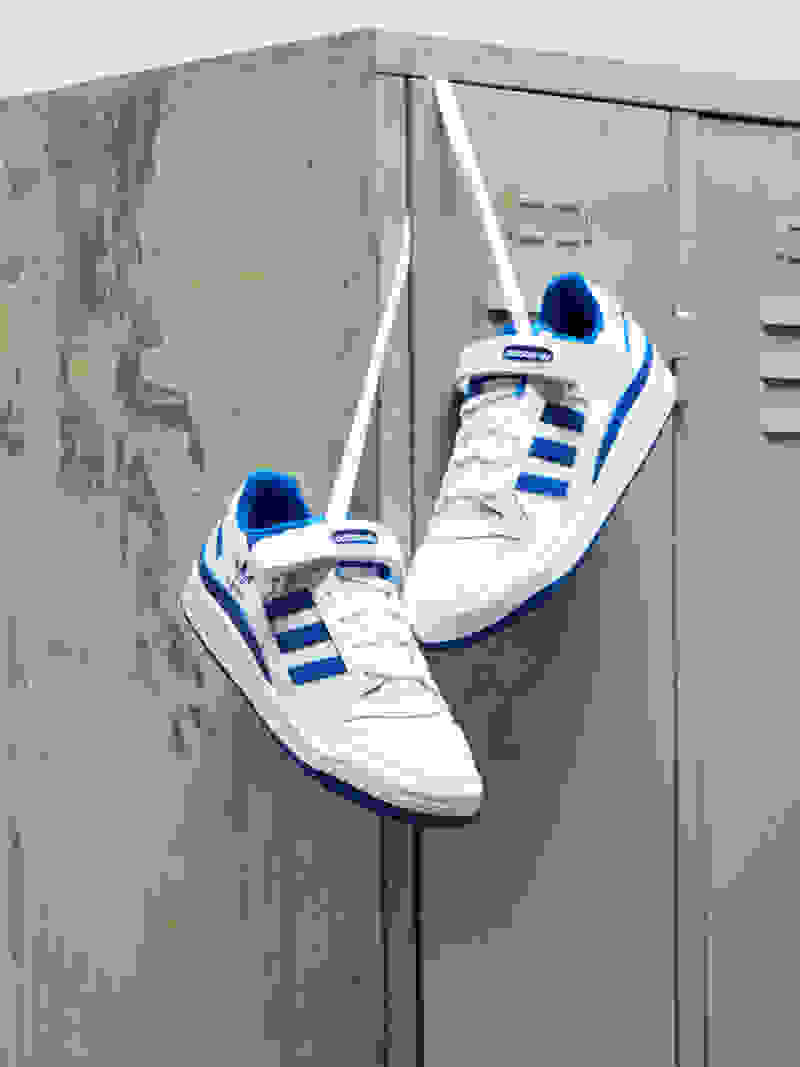 A close-up of a pair of white Forum trainers with blue Three Stripes and accents hanging from their laces on the corner of a metal locker.