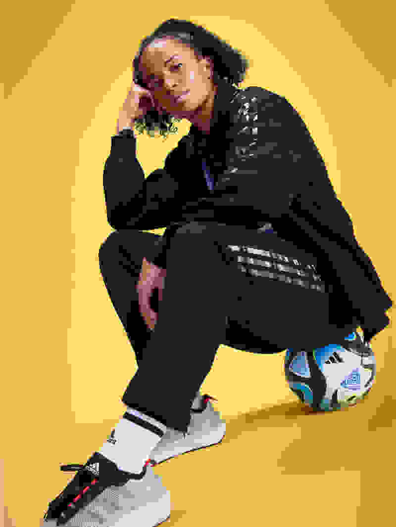 Soccer player Mary Fowler sits posed on a soccer ball, wearing a black adidas tracksuit and sneakers.