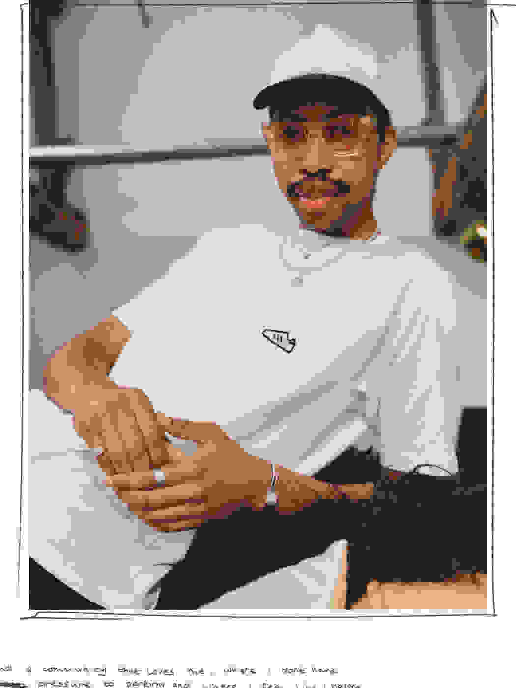 An adiClub member wearing white Stitched Classics white t-shirt and a white cap with black peak, wearing glasses and jewelry, leaning casually on a couch.