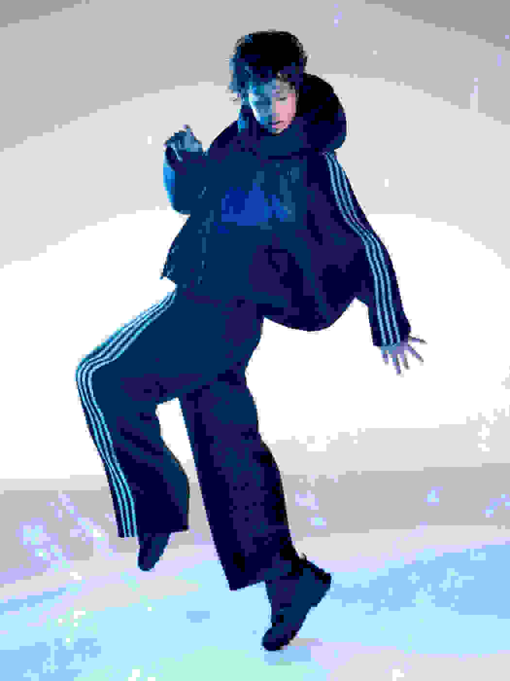 A woman wearing a black 3-striped adidas Y-3 jacket and matching pants jumps high with one leg up while rain falls around her.