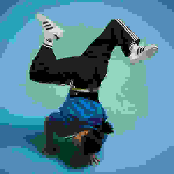 A person in black clothing and white sneakers is performing a handstand against a vibrant teal background.