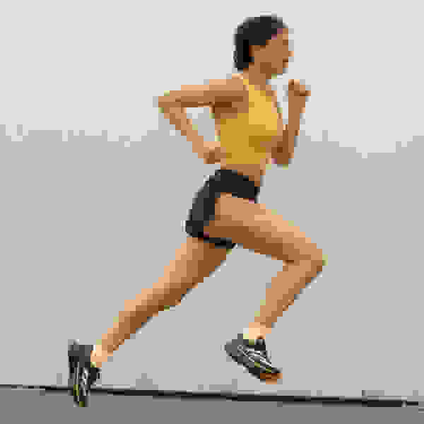 A person in a yellow top and black shorts is running in front of a white wall.