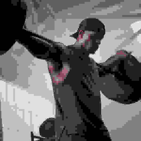 A muscular man in a black shirt and cap is lifting weights in a gym setting with a tiled wall in the background.