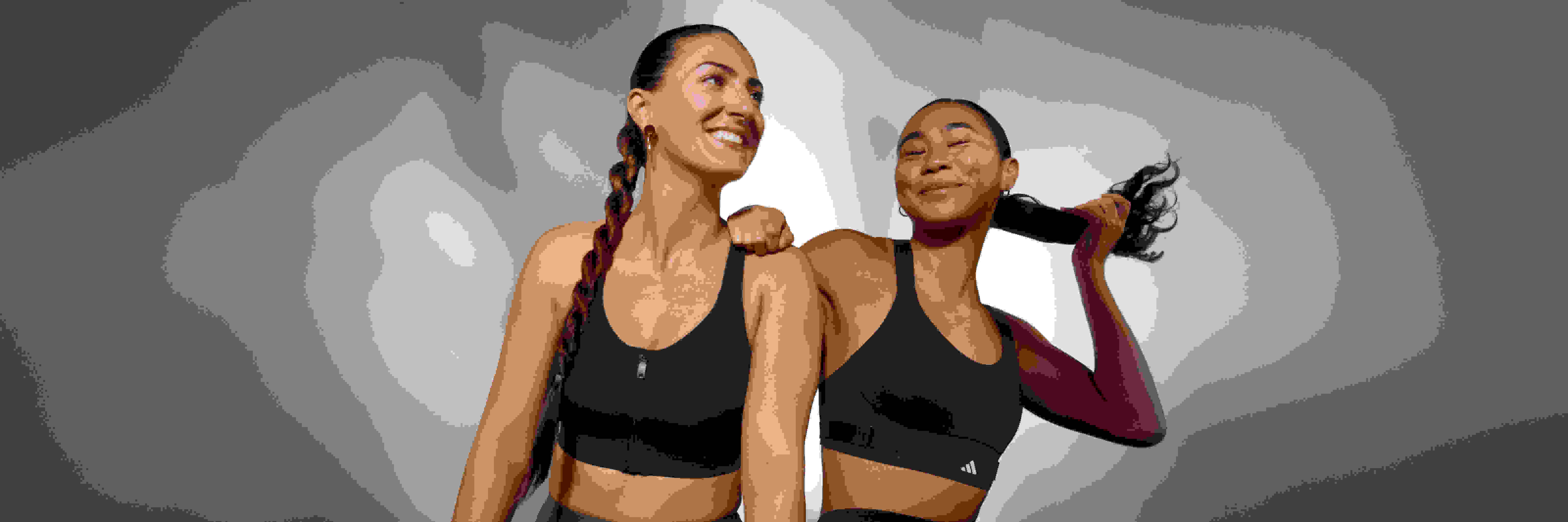 Dos mujeres con sujetadores deportivos negros posando y sonriendo sobre un fondo liso.