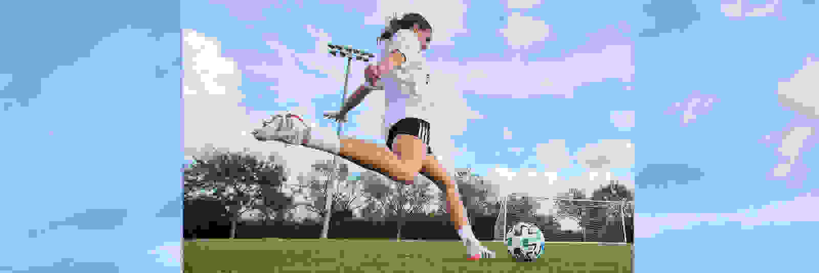 A person in athletic clothing is jumping high in the air on a grassy field, with a cloudy blue sky and trees in the background.