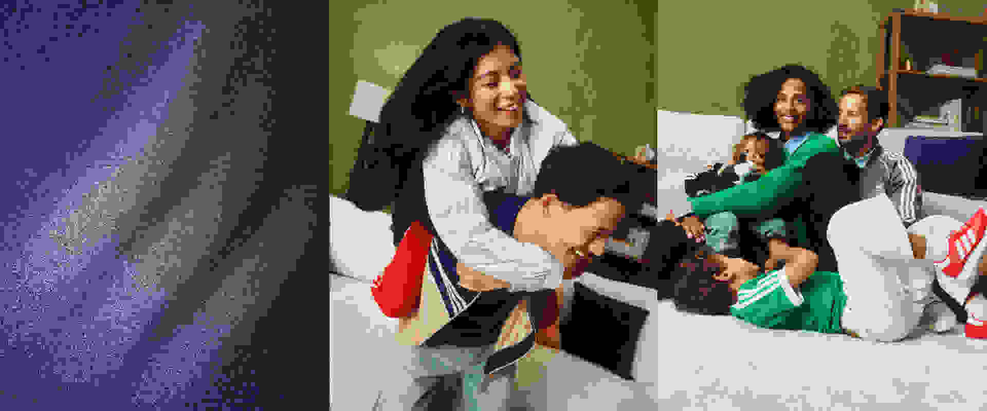 The image shows a woman in a white shirt and red skirt embracing a young child, while a group of people in the background appear to be celebrating or interacting with each other.