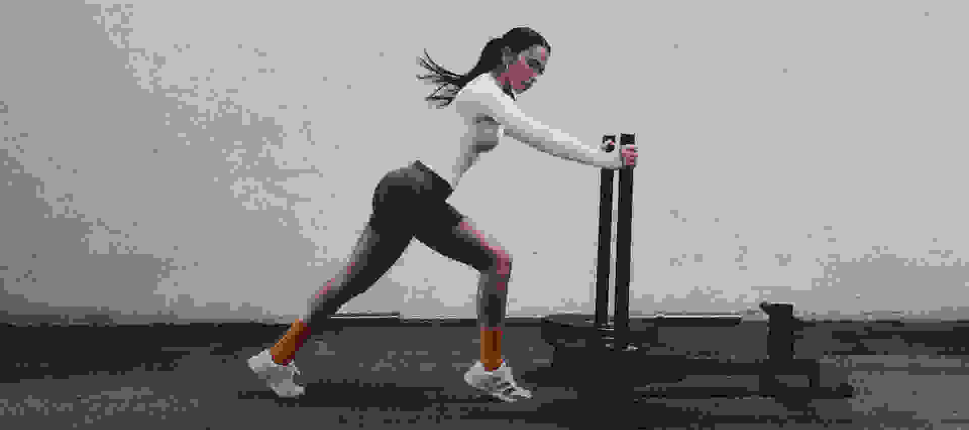 A woman in a white bodysuit and orange sneakers stands in a fitness studio, holding a weight bar and posing against a plain gray wall.