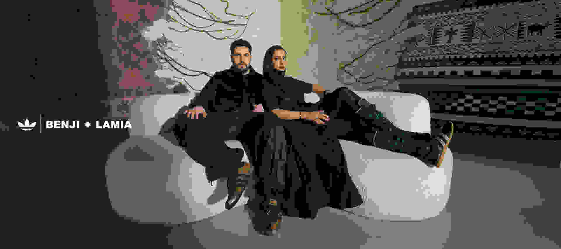 Two individuals, a man and a woman, are seated on a couch against a backdrop of intricate, patterned walls and decorations.