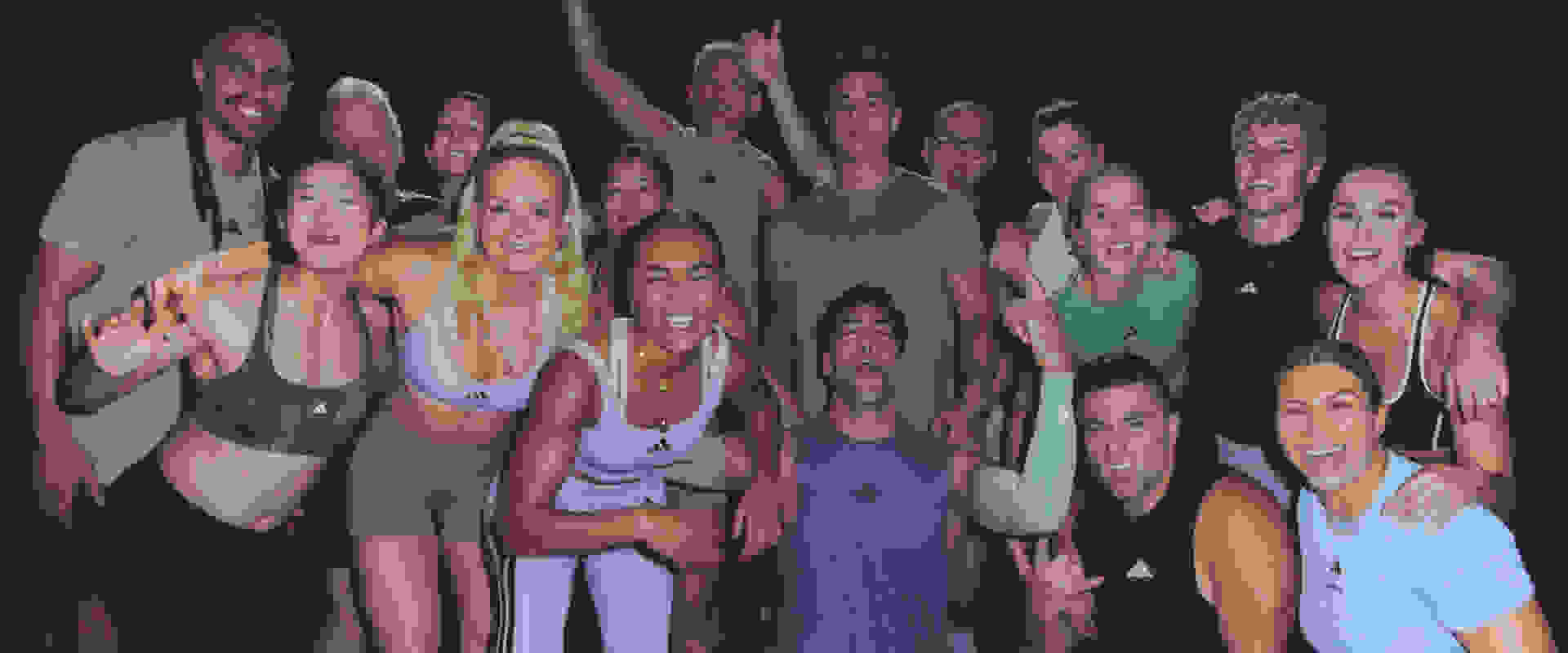 A group of people, mostly women, are gathered together and posing enthusiastically, with some raising their arms in celebration against a dark background.