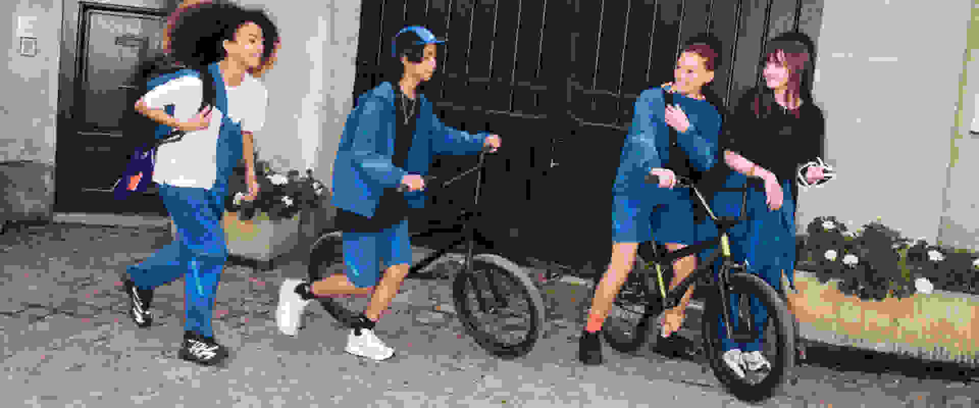 A group of young people, dressed in matching blue outfits, stand together in front of a wooden structure, with one person riding a bicycle.