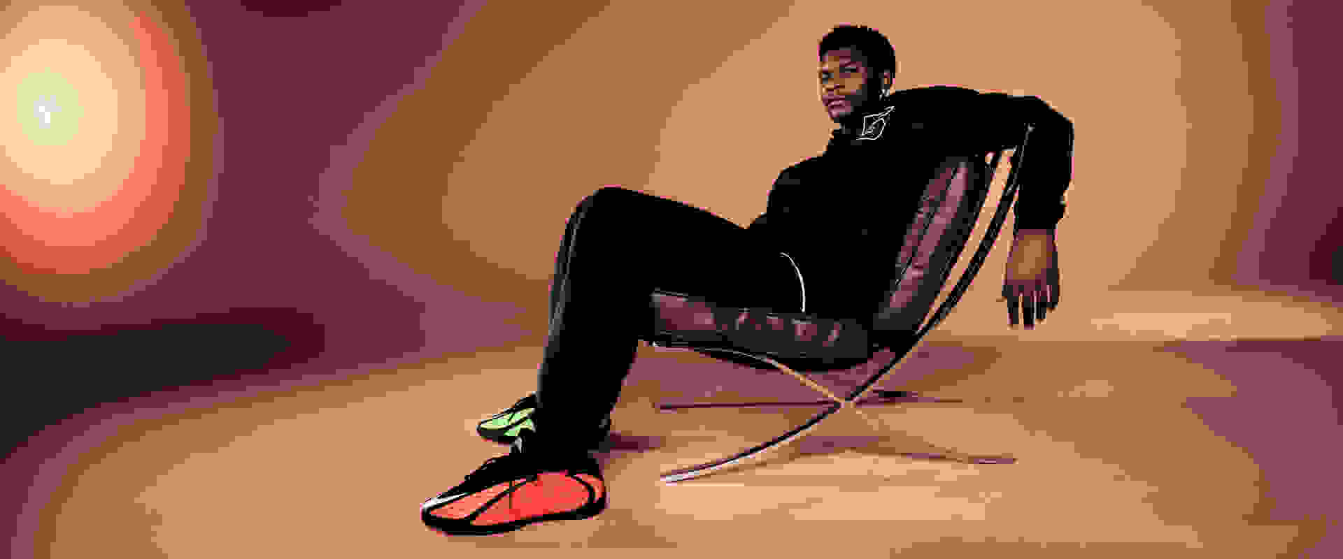 Anthony Edwards portant un survêtement adidas noir assis sur une chaise marron, avec des sneakers noires adidas x Anthony Edwards 2 comportant des détails rouges et verts.