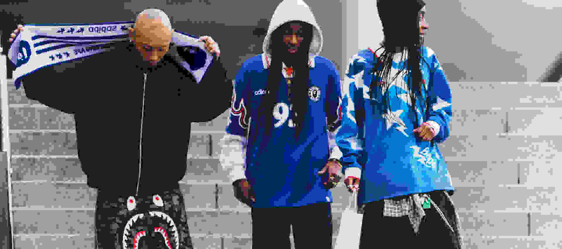 The image shows three individuals wearing hooded jackets, one in black and two in blue, standing in front of a wall with text and graphics.