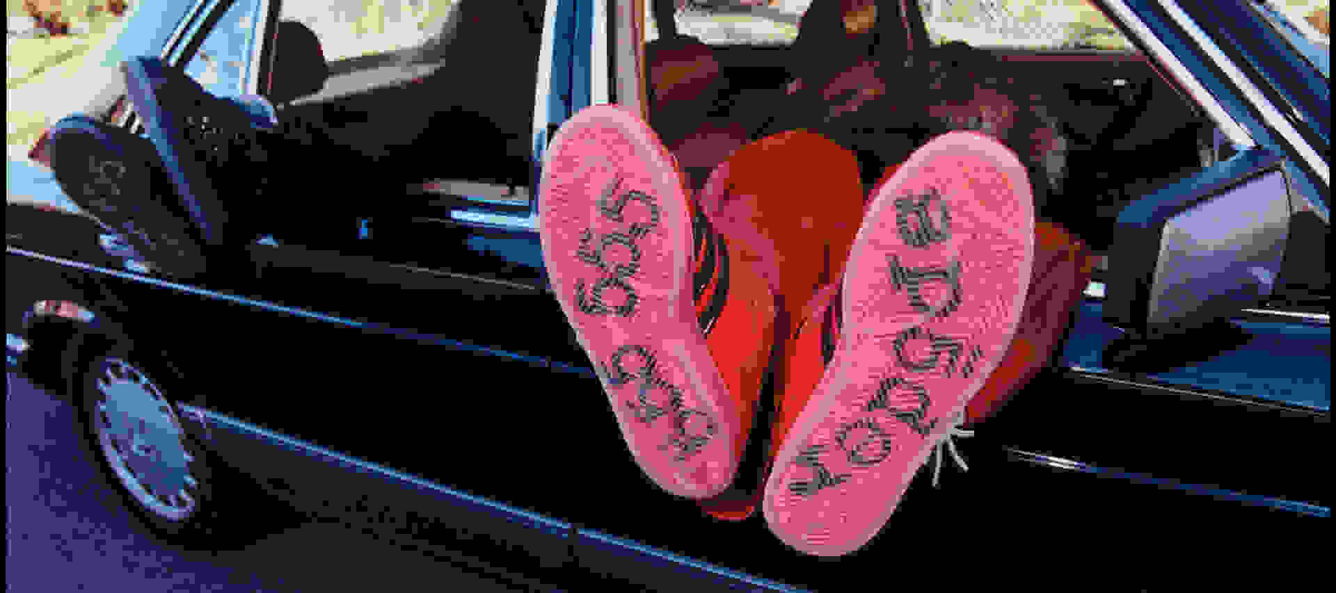 The image shows a pair of bright red flip-flops or sandals placed on the floor of a car, with the car's interior visible in the background.