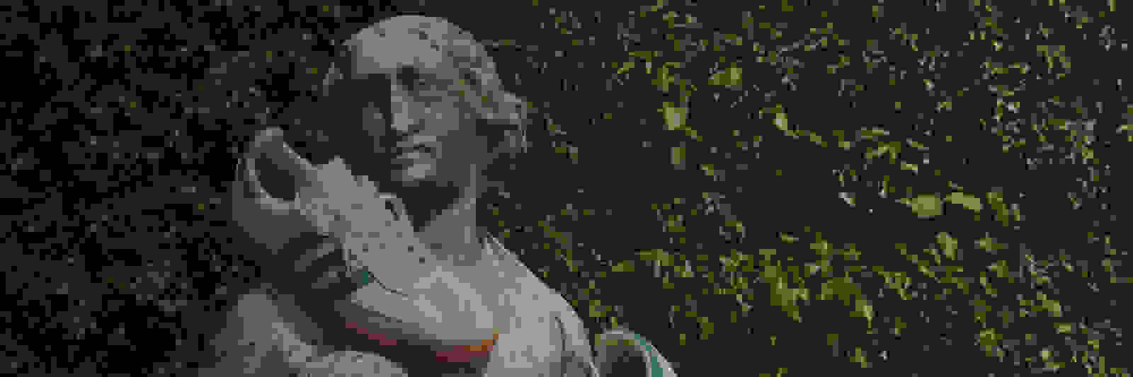 Eine verwitterte Steinstatue einer Figur, die einen Sneaker hält, steht inmitten üppigen, grünen Laubes im Hintergrund.