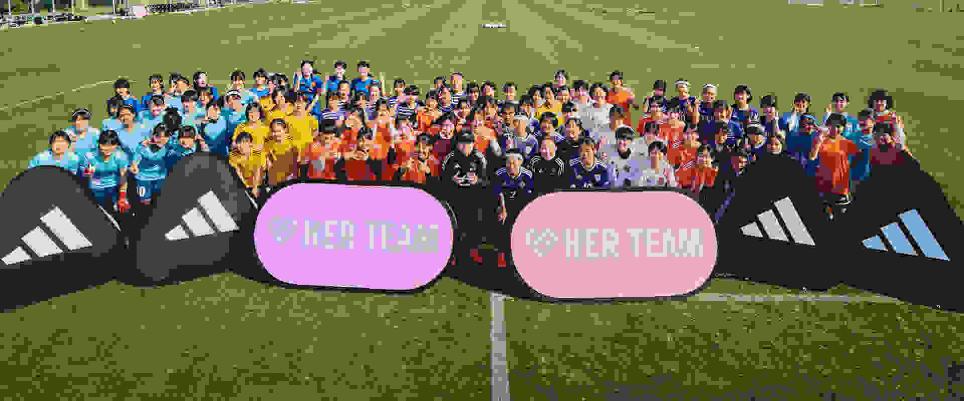 A large group of people wearing colorful jerseys and standing in front of two large pink banners with the text "HER TEAM" on a grassy field.