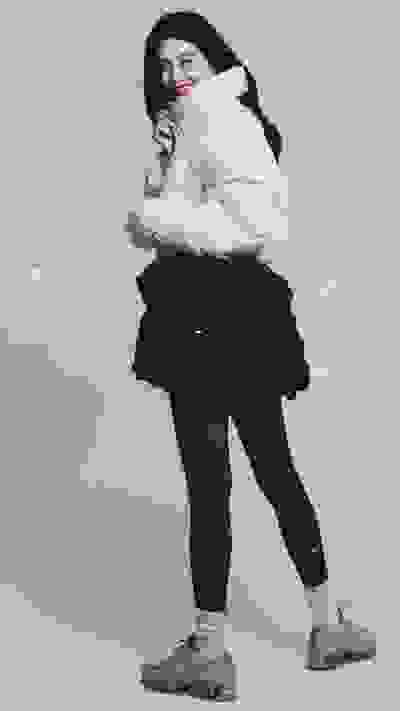 A young woman in a warm winter outfit, smiling and embracing herself, stands in a snowy outdoor setting.