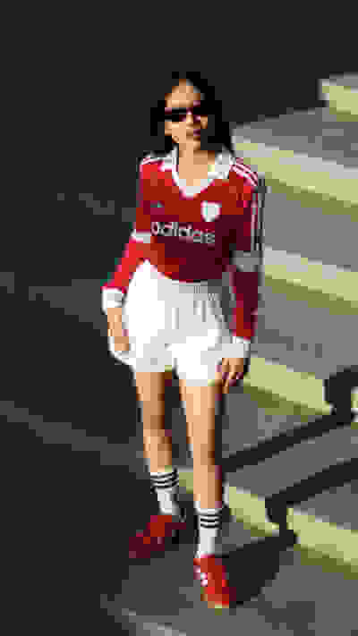 A young woman wearing a red Adidas jersey, white shorts, and red sneakers stands in front of a dark background.