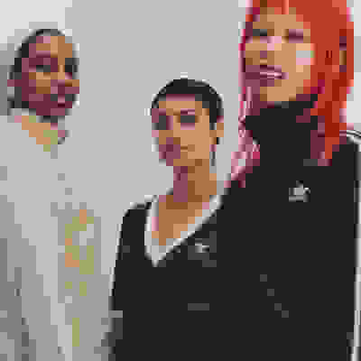 Trois individus, deux femmes et une personne, se tiennent ensemble sur l'image. Ils portent des vêtements casual, comme une veste beige, un haut vert et une veste noire ornée du logo adidas. L'arrière-plan semble être un mur uni de couleur claire.