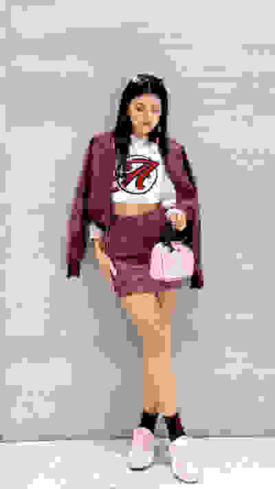A young woman wearing a burgundy jacket, a white crop top, and pink sneakers stands against a plain white wall, holding a small pink purse.