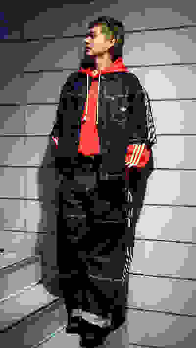 A person wearing a red hoodie, denim jacket, and dark pants stands in front of a tiled wall background.