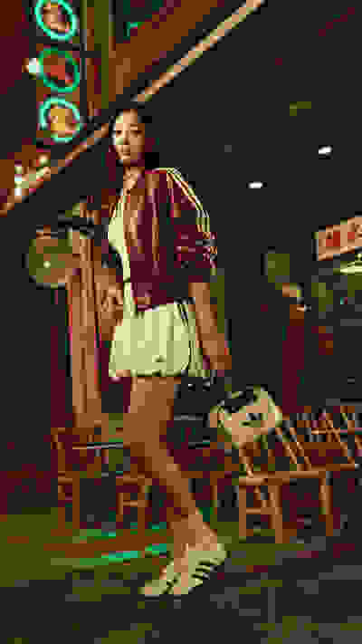 A young woman in a burgundy jacket and white skirt stands in a dimly lit, neon-lit setting with Chinese-style decor in the background.