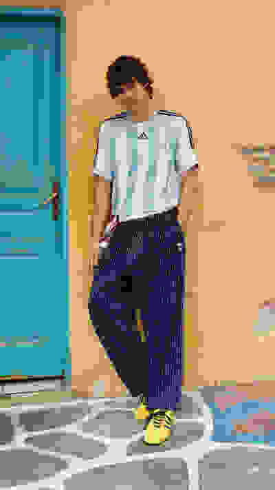 A young person wearing an Adidas jersey and navy blue and white striped pants stands in front of a vibrant turquoise door against a yellow wall.