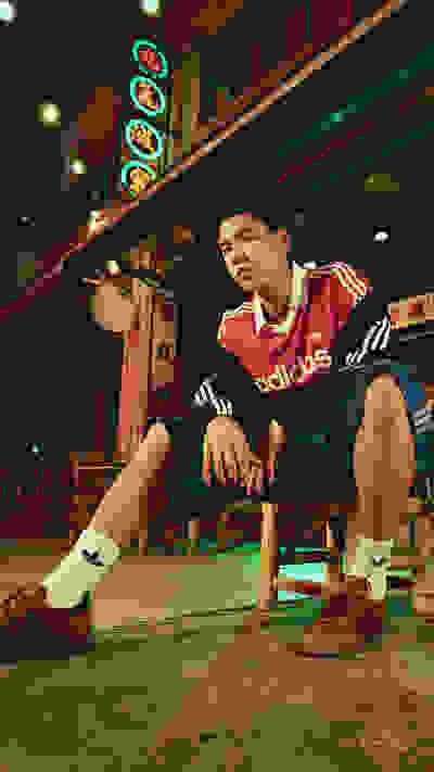 A young man wearing an Adidas tracksuit is sitting on the floor in a dimly lit room with neon signs in the background.
