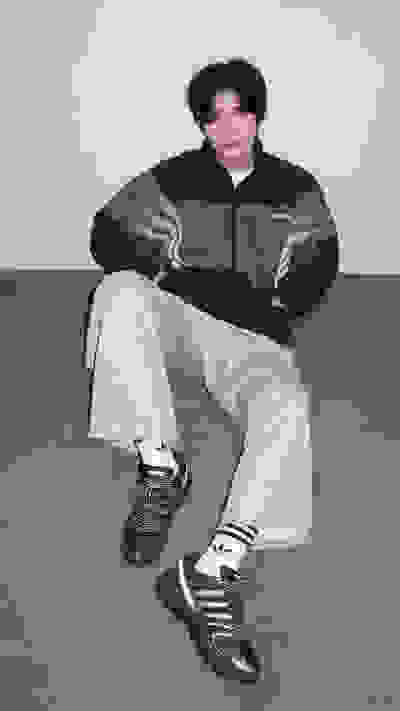 A person wearing a gray and black jacket and light-colored pants is seated on a gray floor, with a plain background behind them.