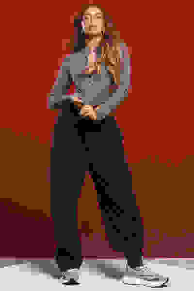 A woman with long, wavy brown hair stands against a vibrant red background, wearing a gray top and black loose-fitting pants, paired with white sneakers.