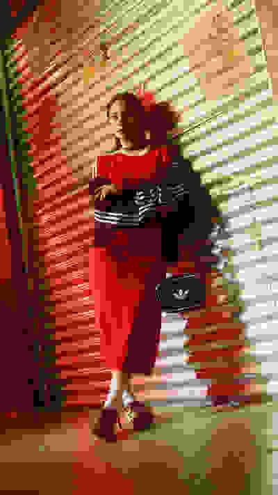 A person wearing a red dress stands in front of a colorful, corrugated metal wall, holding a black handbag.