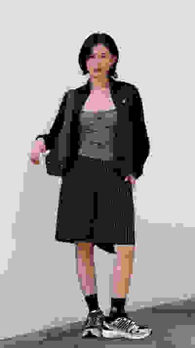 A young woman wearing a black jacket, a patterned top, and striped shorts stands in front of a plain white wall.