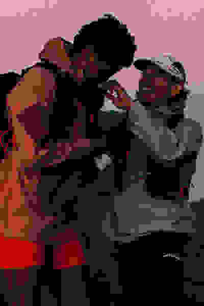 Two people, dressed in warm outdoor clothing, are embracing and smiling at each other against a backdrop of a vibrant sunset sky.