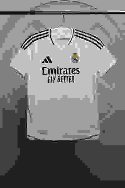 En hvid fodboldtrøje med Real Madrid-logoet og teksten "Emirates fly better" vises på en bøjle med en grå baggrund.