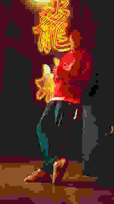 A person in a red sweater sits in front of a vibrant neon sign against a dark background.