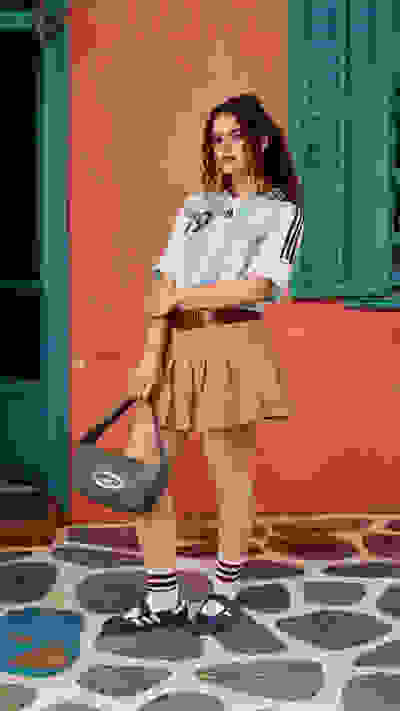 A young woman with curly hair stands in front of a vibrant pink wall, wearing a white sports jersey, beige skirt, and patterned socks with blue sneakers, holding a black and white bag.