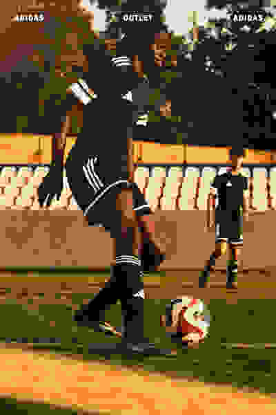 Un futbolista con uniforme de adidas regatea el balón en un campo, con una tienda adidas outlet visible al fondo.