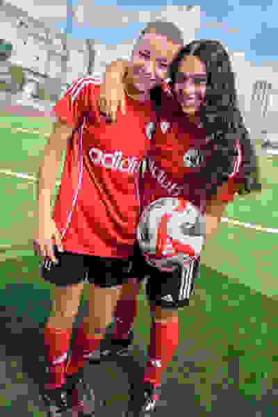 To unge kvindelige fodboldspillere, der bærer røde trøjer, omfavner og smiler på en fodboldbane med en skyet himmel i baggrunden.