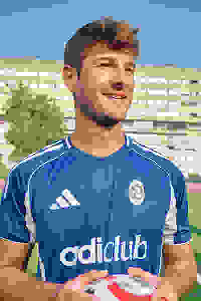 En smilende ung mand, der bærer en blå sportstrøje med logoet "Adiclub", står foran en klods-bygning og holder en fodbold.