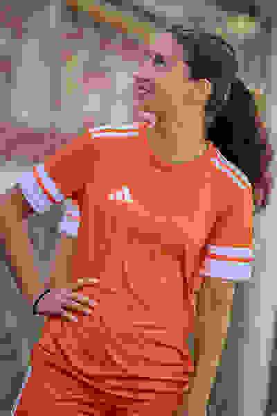 En ung kvinde, der har en orange sportstrøje på, står udenfor og smiler og poserer med hænderne på hofterne på en sløret baggrund.