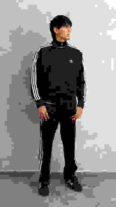A person wearing a black Adidas tracksuit stands in a plain, gray-toned setting.